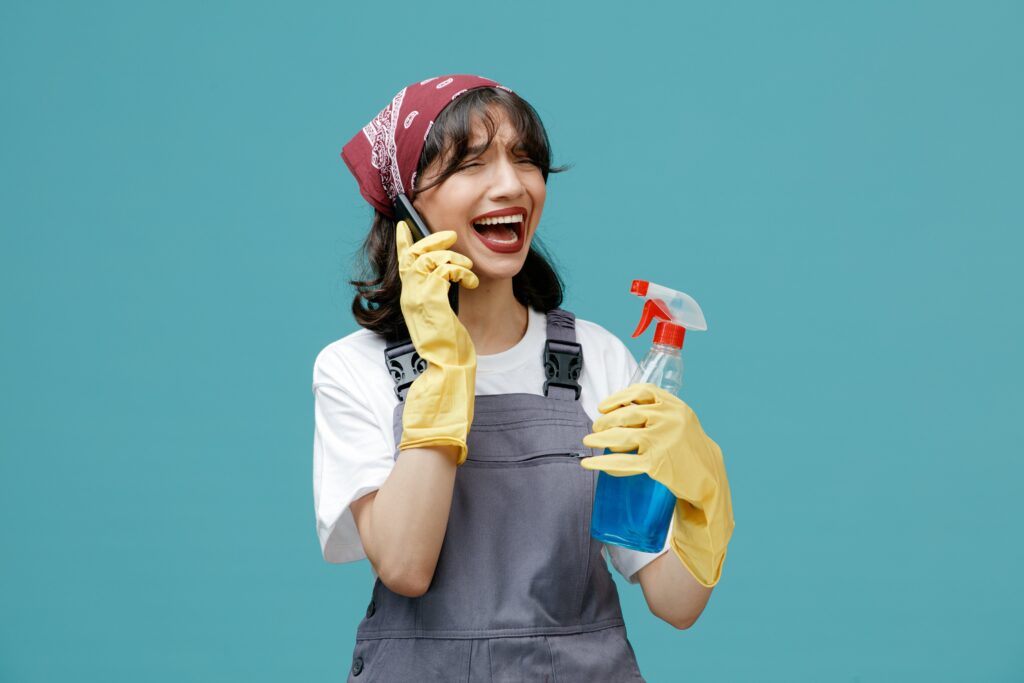 sad-young-female-cleaner-wearing-uniform-bandana-rubber-gloves-holding-cleanser-talking-phone-looking-side-isolated-blue-background-min